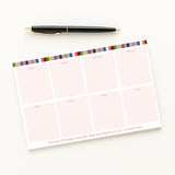 Weekly Notepad with eras colorful stripes at the top and a pen on a white background
